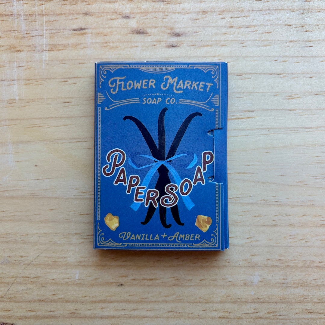 A blue box of Papersoap Vanilla + Amber soap by Flower Market Soap Co. placed on a light wood surface.