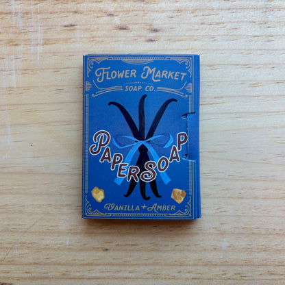 A blue box of Papersoap Vanilla + Amber soap by Flower Market Soap Co. placed on a light wood surface.
