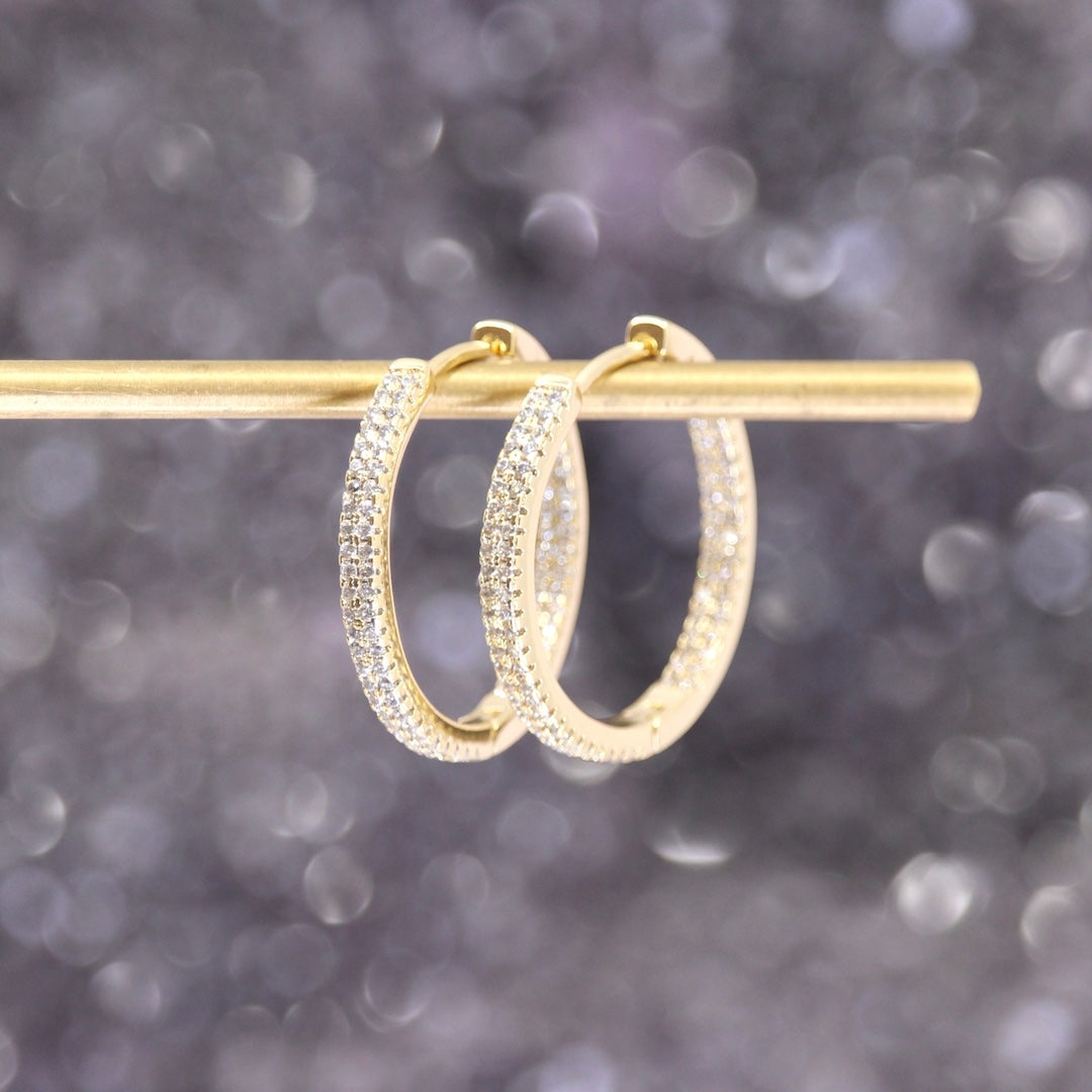A pair of gold hoop earrings with pavé-set clear stones, hanging from a gold display rod against a soft, out-of-focus purple and gray background.