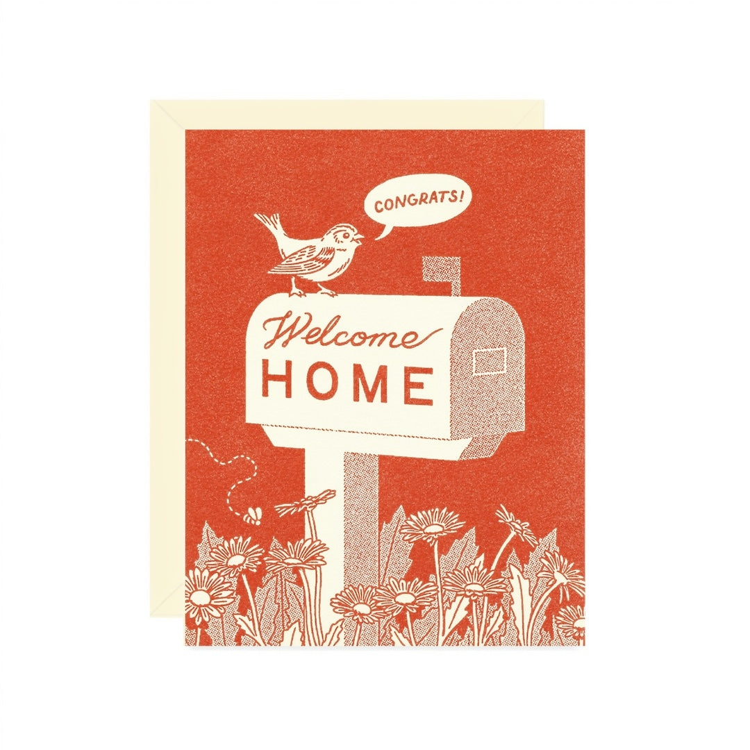 A greeting card with a red-orange background and cream illustration of a bird on a mailbox that says “Welcome Home,” with a speech bubble reading “Congrats!” A cream envelope is partially visible behind the card on a white background.