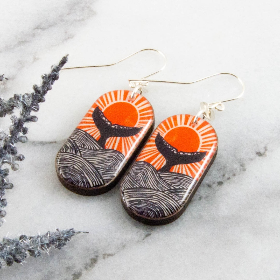 Pair of earrings with a whale tail and sun design on a marble surface