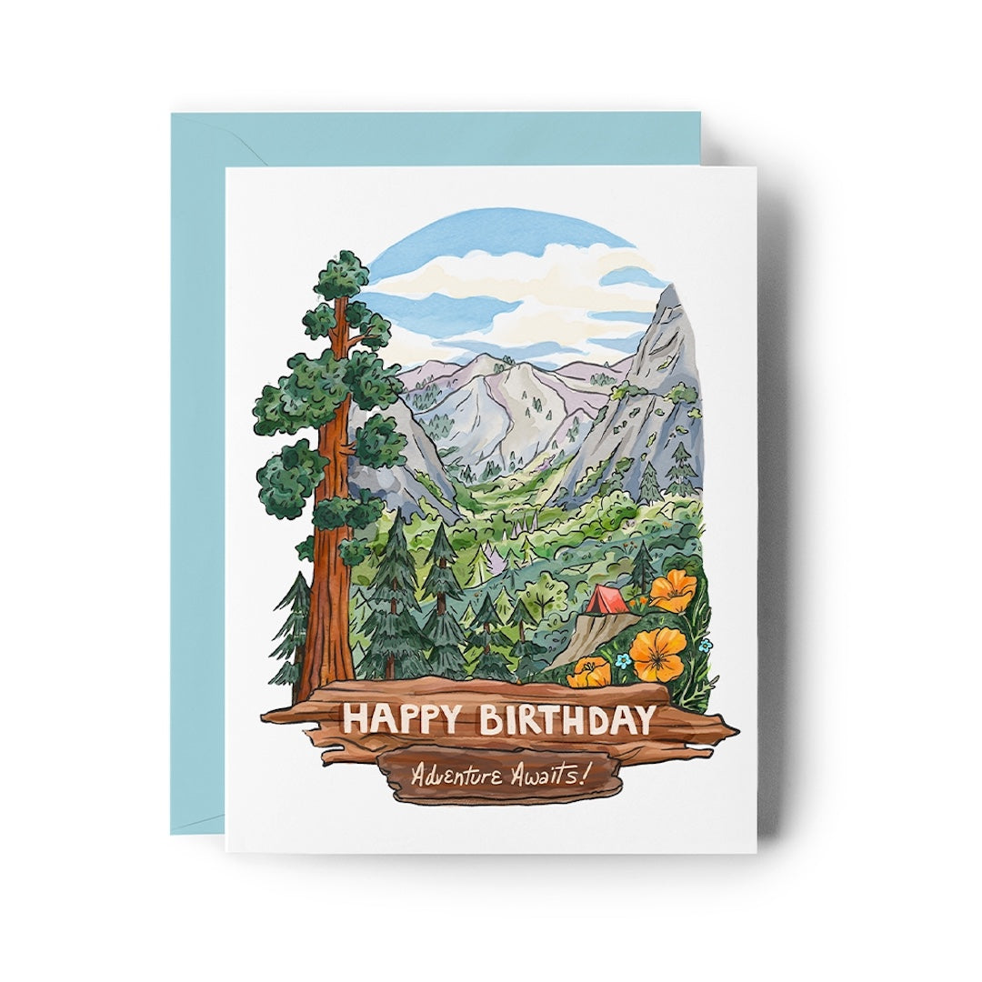 A square birthday card with a scenic mountain landscape illustration, shown with a light blue envelope behind it on a white background. The card reads “HAPPY BIRTHDAY” and “Adventure Awaits!” on a wooden sign graphic at the bottom.