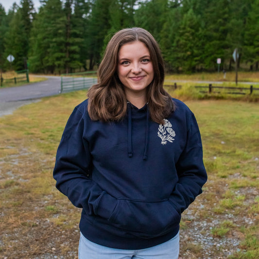 A woman with brown hair stands outdoors wearing a navy blue hoodie with a white floral graphic on the left chest, paired with light blue jeans. The background shows a grassy field, a dirt path, and a forest of green trees.