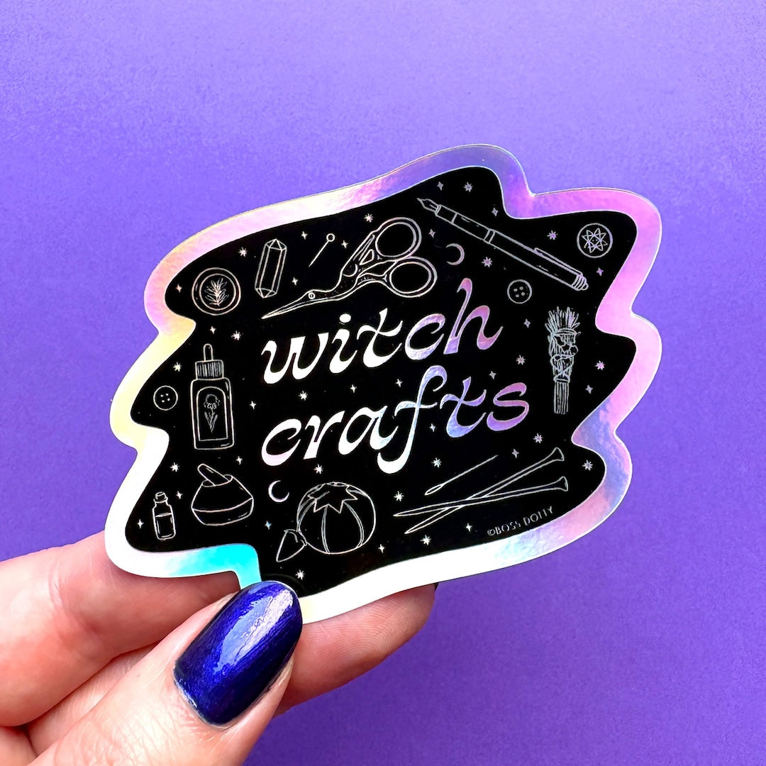 A hand with dark blue nail polish holds a die-cut sticker featuring a black background with white witchcraft and regular craft illustrations and the words 'witch crafts' in white script with a purple outline. The sticker has an iridescent holographic border and is set against a solid purple background.