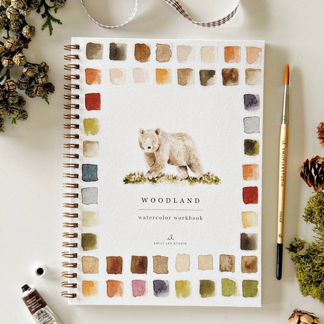 Watercolor workbook with bear illustration and 'Woodland' text on a white surface with stationery items.