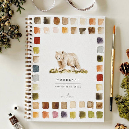 Watercolor workbook with bear illustration and 'Woodland' text on a white surface with stationery items.