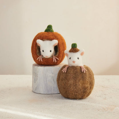 Two felted wool mouse figurines peeking from pumpkins, displayed on a light surface against a soft beige background.