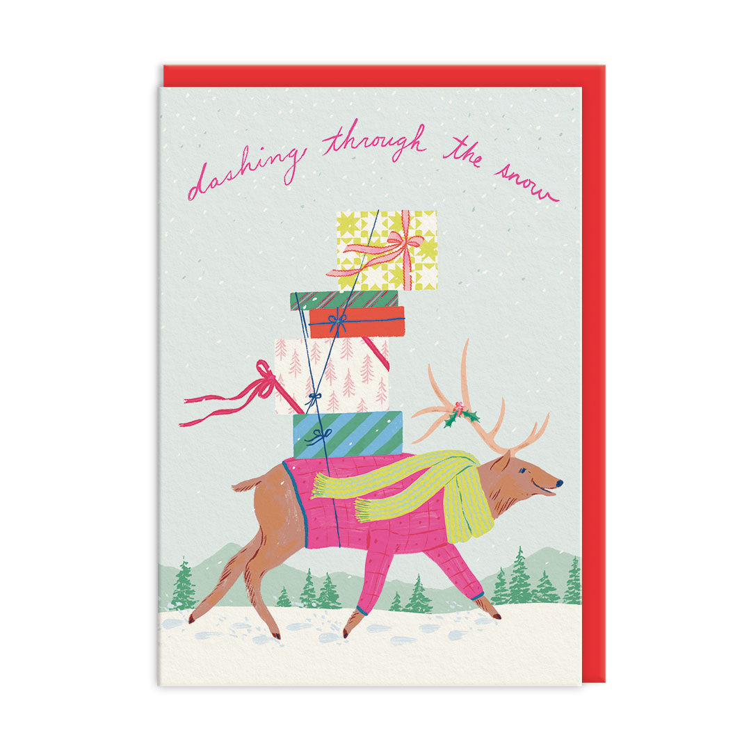 A Christmas greeting card featuring an illustrated reindeer carrying wrapped gifts while walking through the snow, with the phrase 'dashing through the snow' in pink script at the top. The card is shown with a red envelope against a white background.