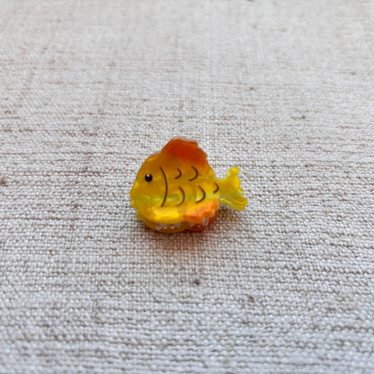 A small yellow and orange fish-shaped hair clip with black scale details, placed on a textured beige fabric background.