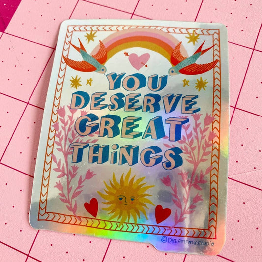 Holographic sticker with motivational text 'You deserve great things' on a pink grid background